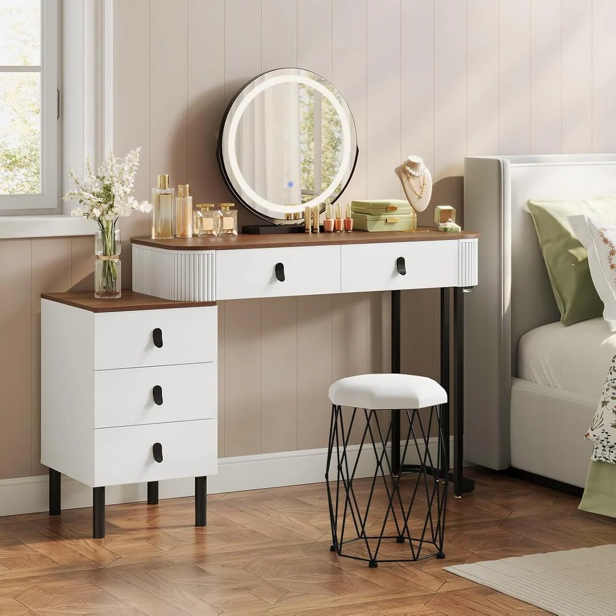 Wood and white dressing table in a bedroom, with its round mirror lit up, drawer unit and velvet stool